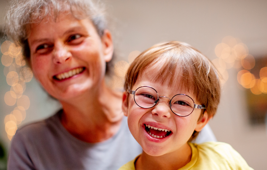 Eine Frau mit grauem Haar hat einen kleiner blonden Jungen mit Brille auf ihrem Schoß. Beide lachen herzlich in die Kamera.