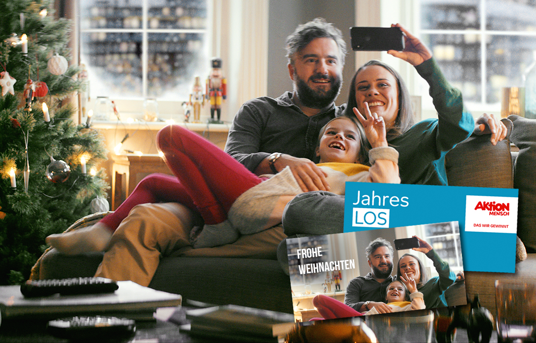 Eine Familie sitzt ein beieinander auf einem Sofa im weihnachtlichen Wohnzimmer. Die Eltern und ihre Tochter nehmen gerade ein gemeinsames Selfie auf. Im Vordergrund ist ein Jahreslos zu sehen, das mit dem Selfie gestaltet ist.