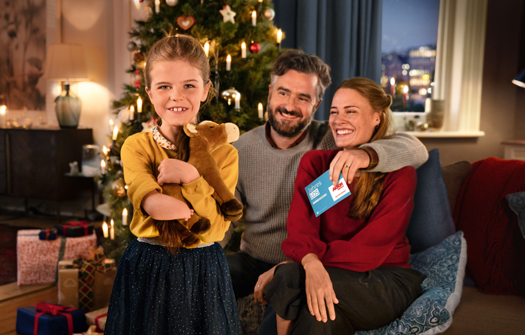 Eine Familie im weihnachtlich geschmückten Wohnzimmer. Die Eltern sitzen lächelnd Arm in Arm auf dem Sofa. Ihre Tochter steht davor und hält glücklich ein Stoffpony im Arm. 