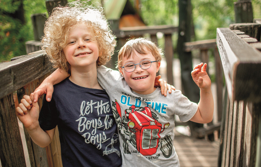 Ein Junge mit Downsyndrom hat seinem Freund ohne Behinderung den Arm kumpelhaft um die Schulter gelegt. Beide lächeln in die Kamera.