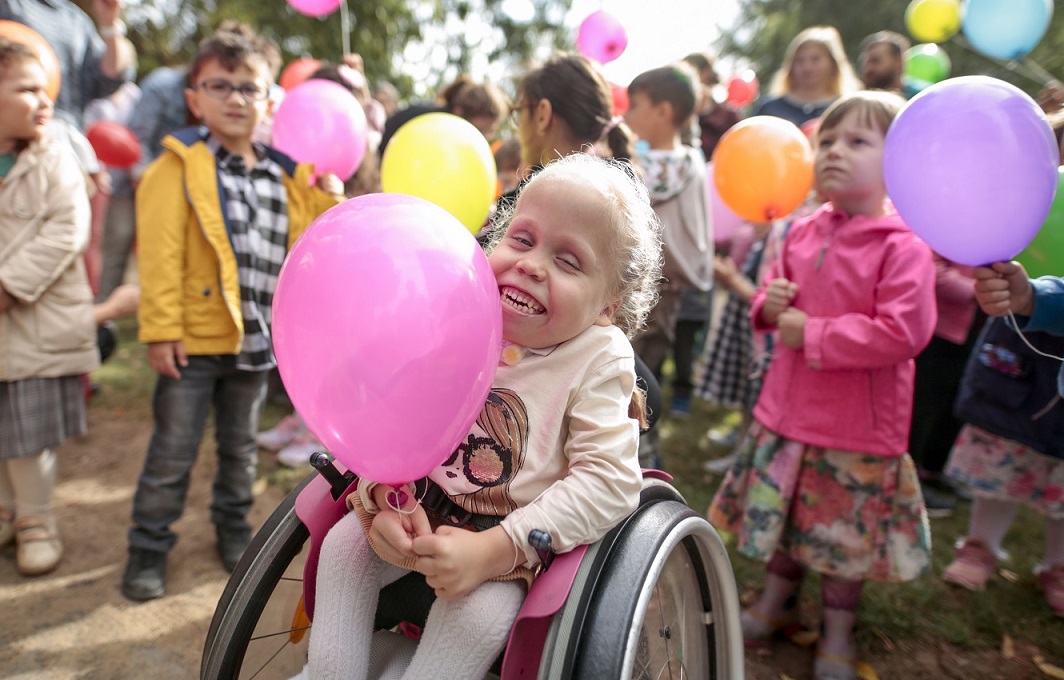 Ein Mädchen im Rollstuhl sitzt inmitten vieler anderer Kinder. Sie halten alle bunte Luftballons in den Händen und feiern ein Fest.