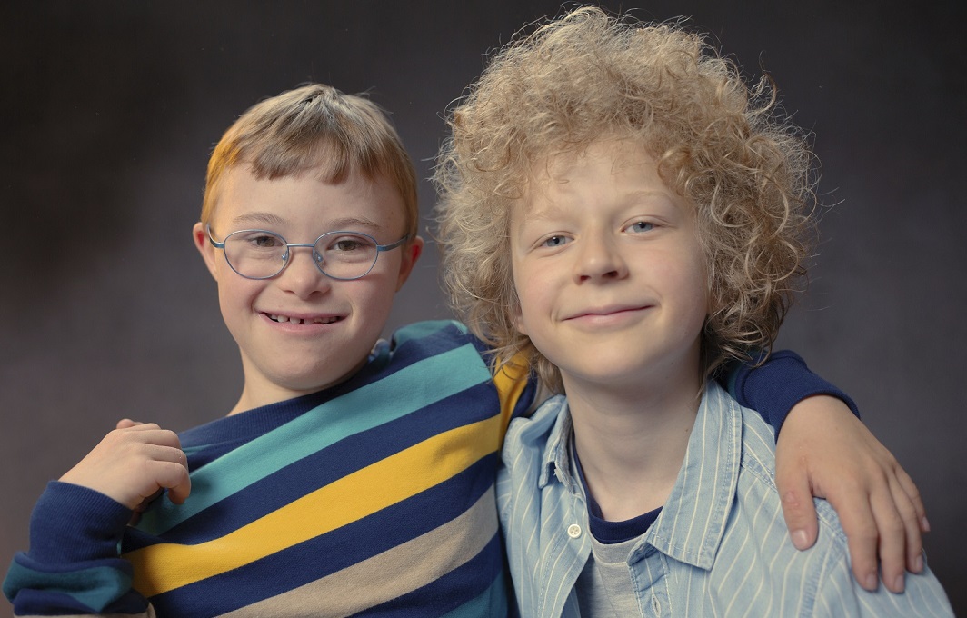 Ein Junge mit Downsyndrom hat seinem Freund ohne Behinderung den Arm kumpelhaft um die Schulter gelegt. Beide lächeln in die Kamera.