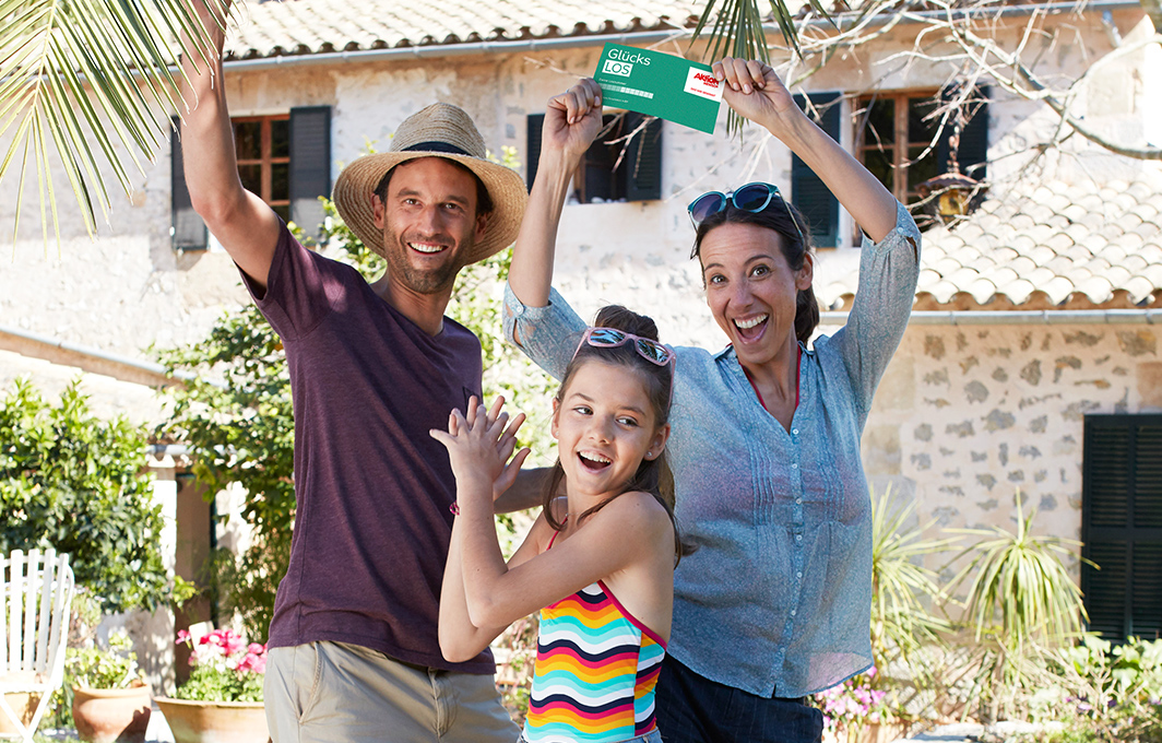 Familie aus Mutter, Vater und Tochter jubeln vor eine Finca mit einem Glückslos in der Hand