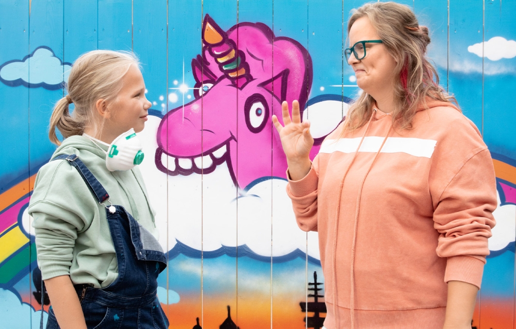 Ein Mädchen und eine Frau stehen sich einander zugewandt gegenüber. Im Hintergrund ist ein buntes Graffiti mit einem Einhorn zu sehen. 