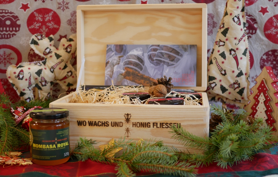 Eine kleine Holzkiste mit der Aufschrift: Wo Wachs und Honig fließen. Sie steht inmitten einer weihnachtlichen Dekoration und enthält mehrere Honiggläser.