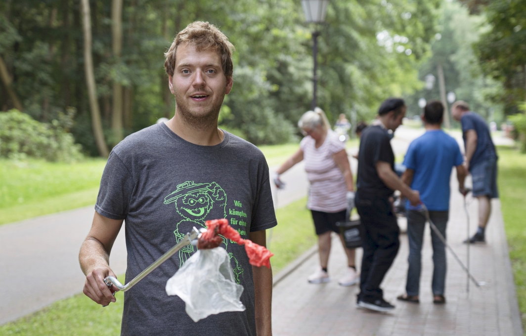 Jan Kleen steht in einem Park und hält eine Müllzange in der Hand. Im Hintergrund sind weitere Müllsammler*innen zu sehen.
