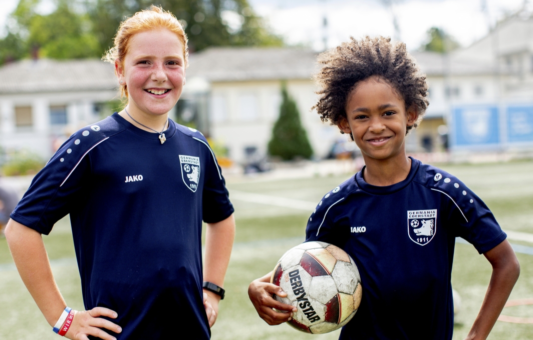 Ein Mädchen und ein Junge stehen nebeneinander in Trikots auf dem Fußballplatz.