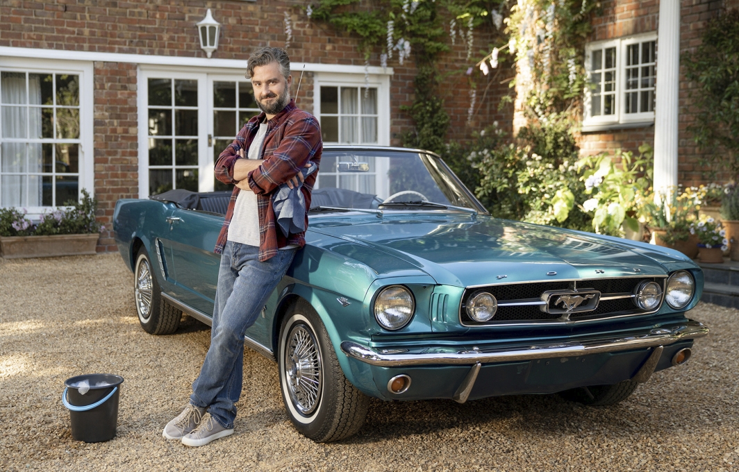 Ein Mann lehnt an einem petrolfarbenen Ford Mustang Cabrio. 