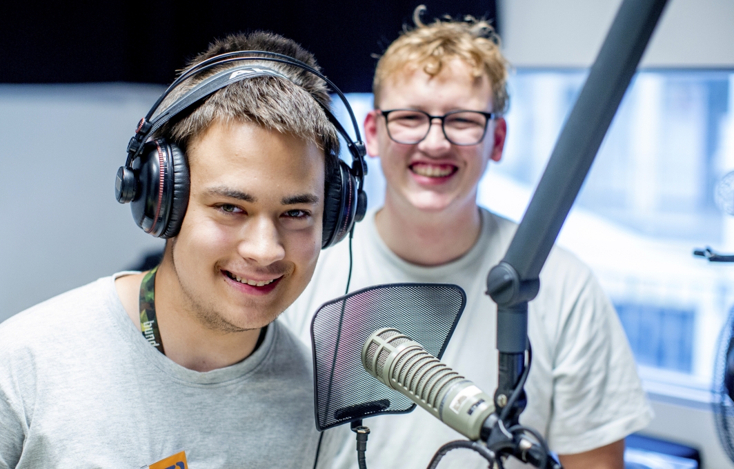 Zwei junge Männer im Aufnahmestudio. Einer von beiden trägt Kopfhörer und steht vor einem Mikrofon.