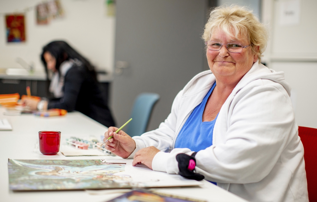 Eine Frau mit blonden Haaren und Brille sitzt an einem Tisch. Sie hält einen Pinsel in der Hand und lächelt in die Kamera. Vor ihr liegt ein halbfertiges Bild.