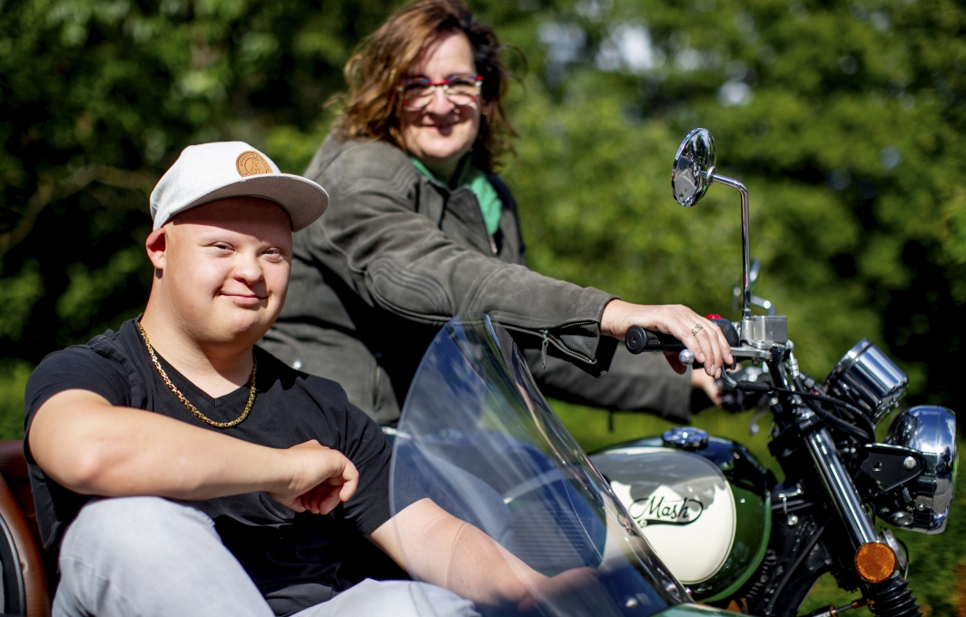 Ein junger Mann mit Downsyndrom und eine Frau sitzen gemeinsam auf einem Motorrad.