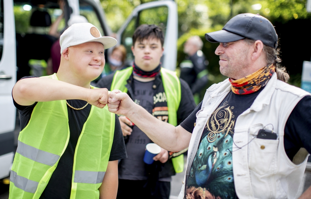 Ein junger Mann mit Downsyndrom und ein älterer Mann sehen sich freundschaftlich an und begrüßen sich mit einem Faustgruß.