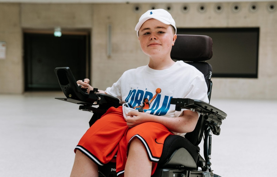 Carl-Josef sitzt in einem E-Rollstuhl. Er trägt eine Baseball-Kappe und sportliche Kleidung und lacht in die Kamera.
