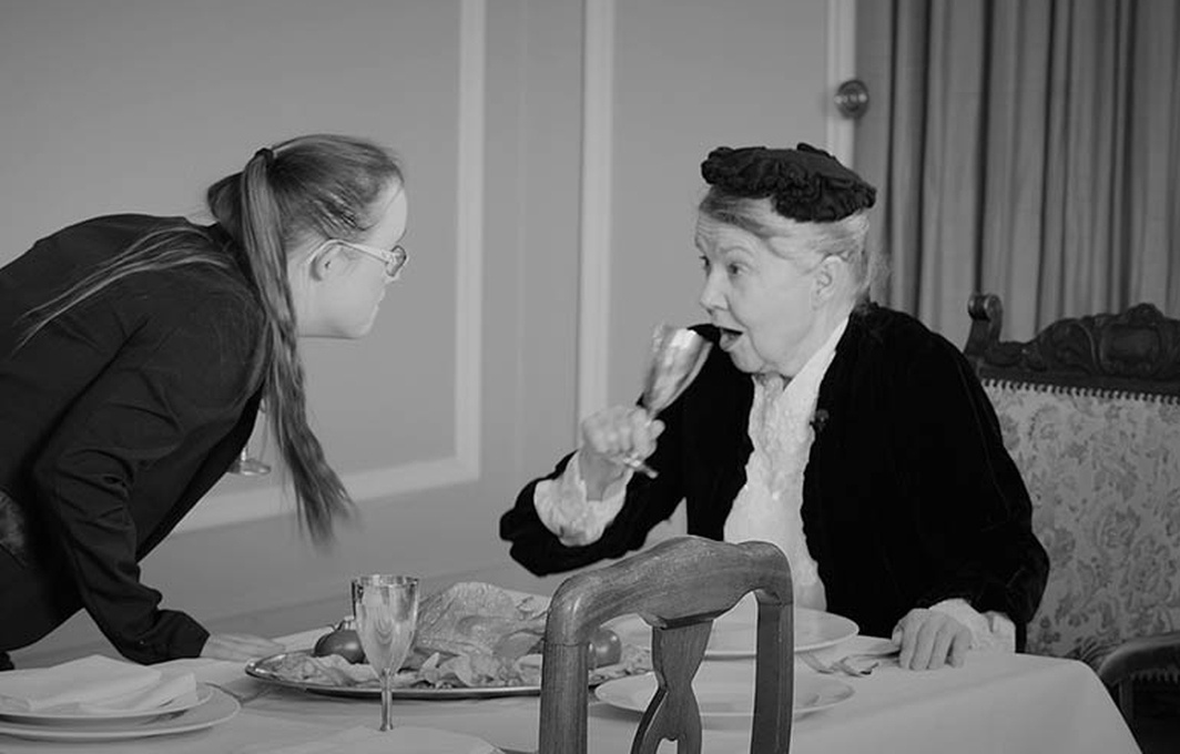 Eine ältere Dame sitzt an einem fein gedeckten Tisch. Ihr gegenüber steht eine junge Frau mit Downsyndrom in Butler-Kleidung. Sie hat sich weit vorgebeugt und schaut der älteren Dame direkt ins Gesicht.