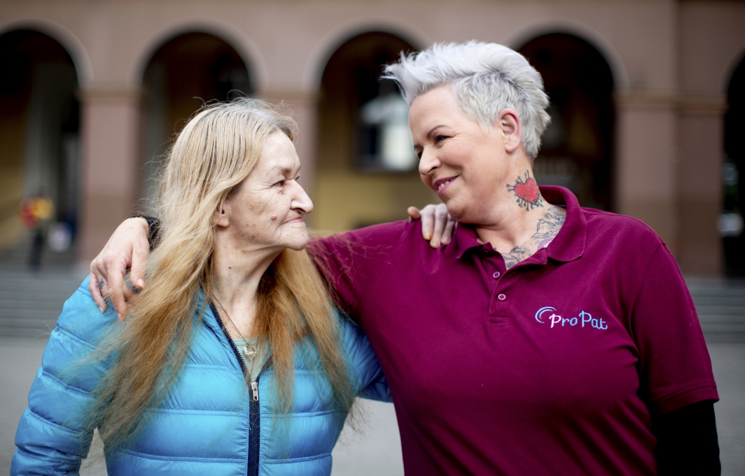 Zwei Frauen stehen Arm in Arm nebeneinander und lächeln in die Kamera. Die eine trägt schulterlanges Haar, die andere einen Kurzhaarschnitt und Tätowierungen am Hals.