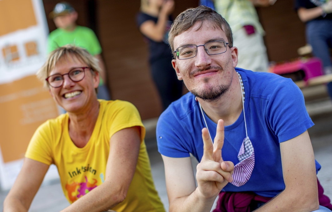 Ein Mann und eine Frau sitzen draußen nebeneinander vor einer Hütte. Er zeigt das Siegeszeichen in die Kamera, beide lächeln.
