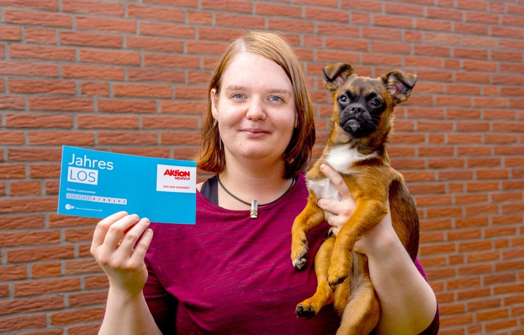 Eine junge Frau mit halblangen Haaren lächelt in die Kamera. Sie hält einen kleinen Hund im Arm und in der einen Hand ein Jahres-Los.