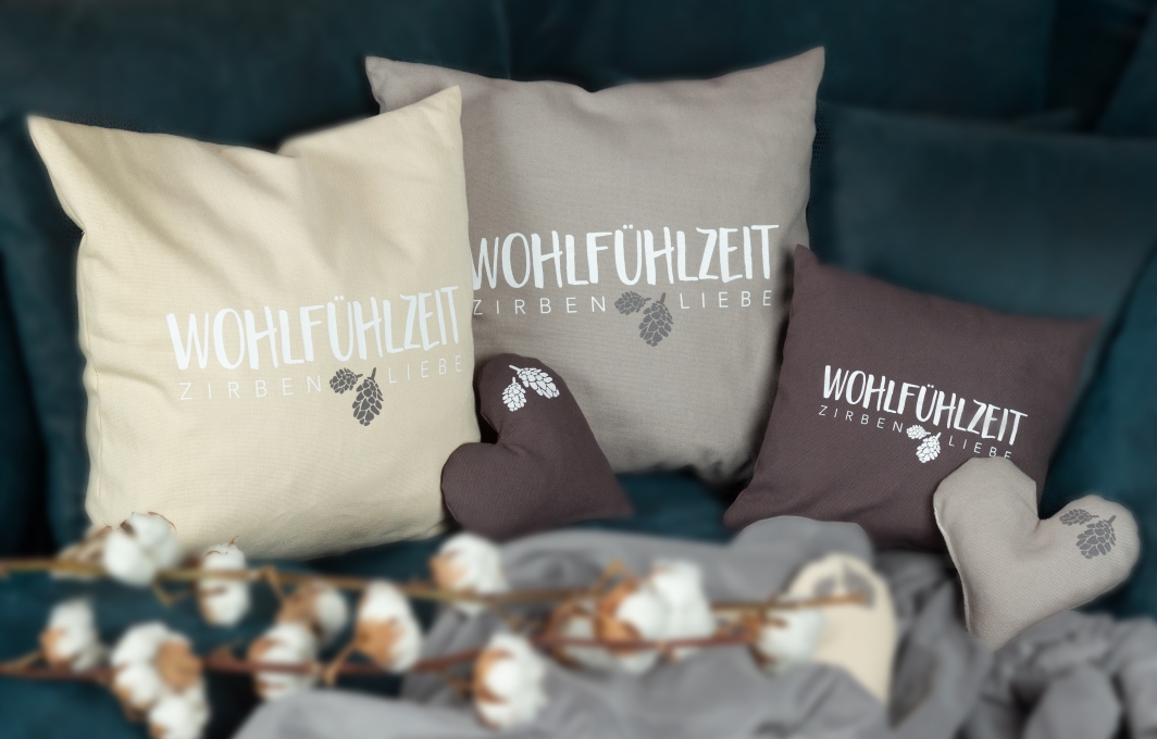 Einige Kissen mit der Aufschrift „Wohlfühlzeit. Zirben-Liebe“ sind nebeneinander drapiert.
