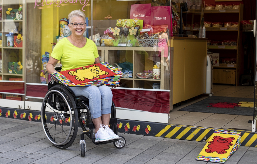 Eine Frau sitzt im Rollstuhl vor einem Geschäft. Sie präsentiert eine Rampe aus Lego. Vor dem Eingangstufe zum Laden liegt ebenfalls eine Legorampe.