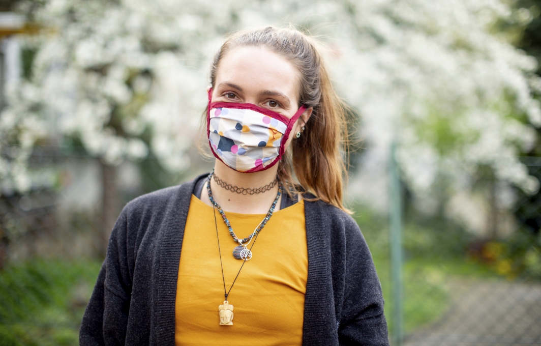 Eine junge Frau mit Pferdeschwanz. Sie trägt eine bunt gemusterte Mund-Nasen-Bedeckung und schaut direkt in die Kamera.