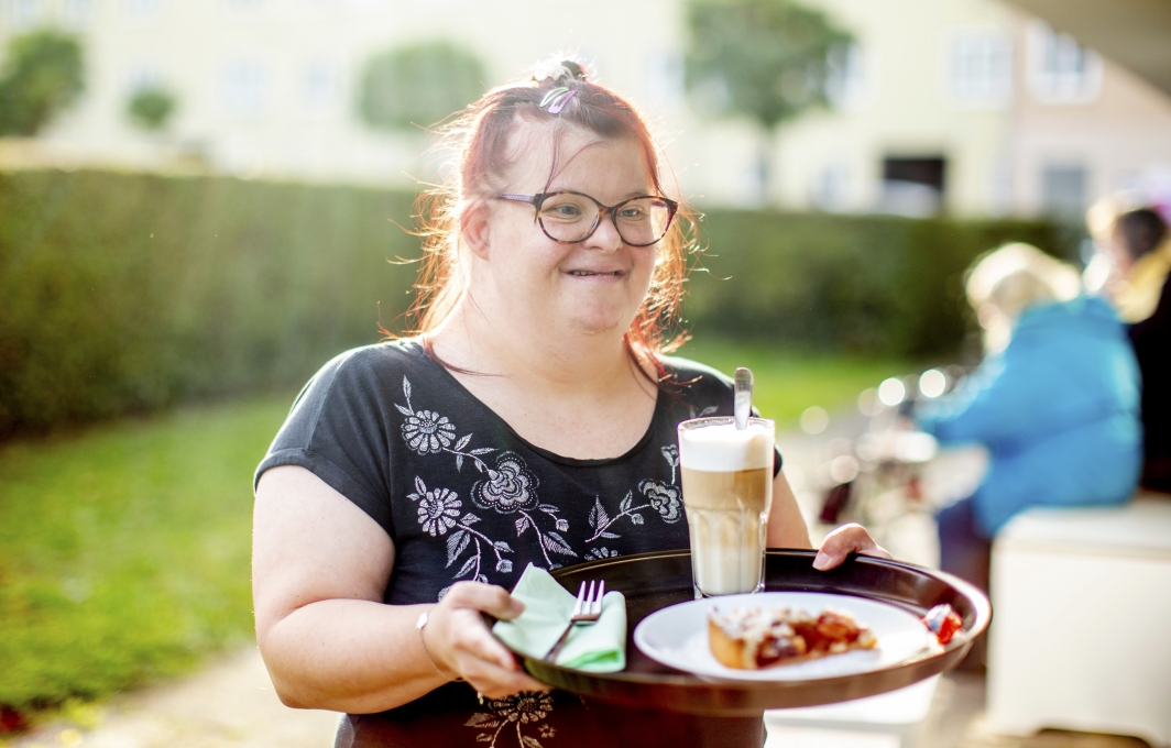 Eine Frau mit Down-Syndrom bedient CafÃ©-Gäste. Sie trägt ein Tablett mit einem Stück Kuchen und Kaffee.