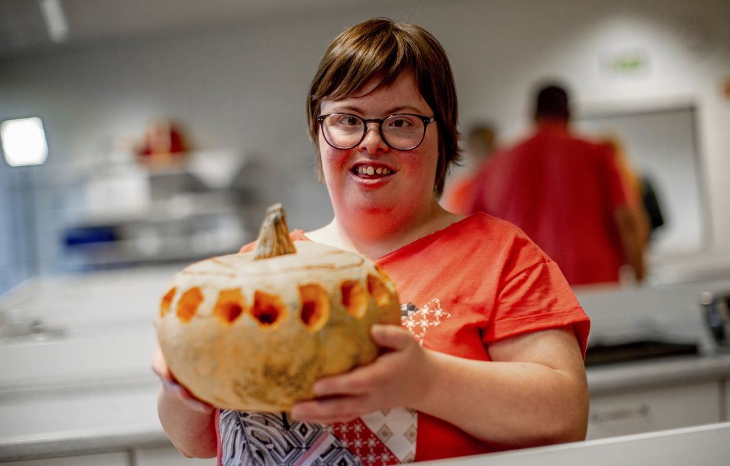 Eine junge Frau mit Down-Syndrom hält einen selbstgeschnitzten Kürbis in den Händen. Sie trägt eine Brille und lächelt in die Kamera.