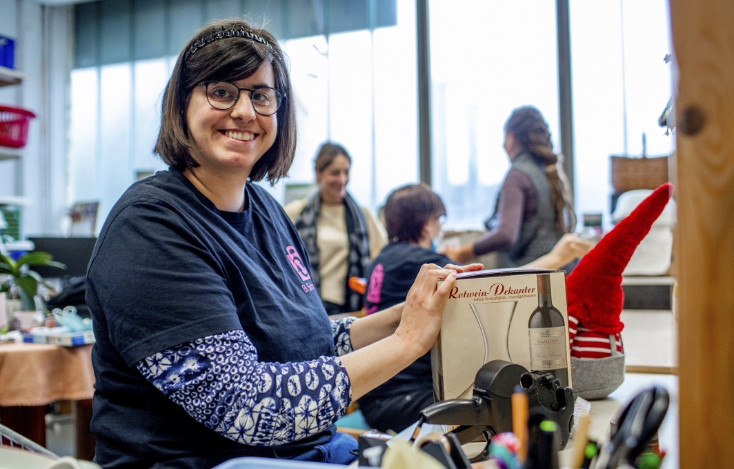 Eine Frau mit Brille und dunklen Haaren sortiert Waren für das Second-Hand-Warenhaus. Sie sitzt an einem Tisch und lächelt in die Kamera.