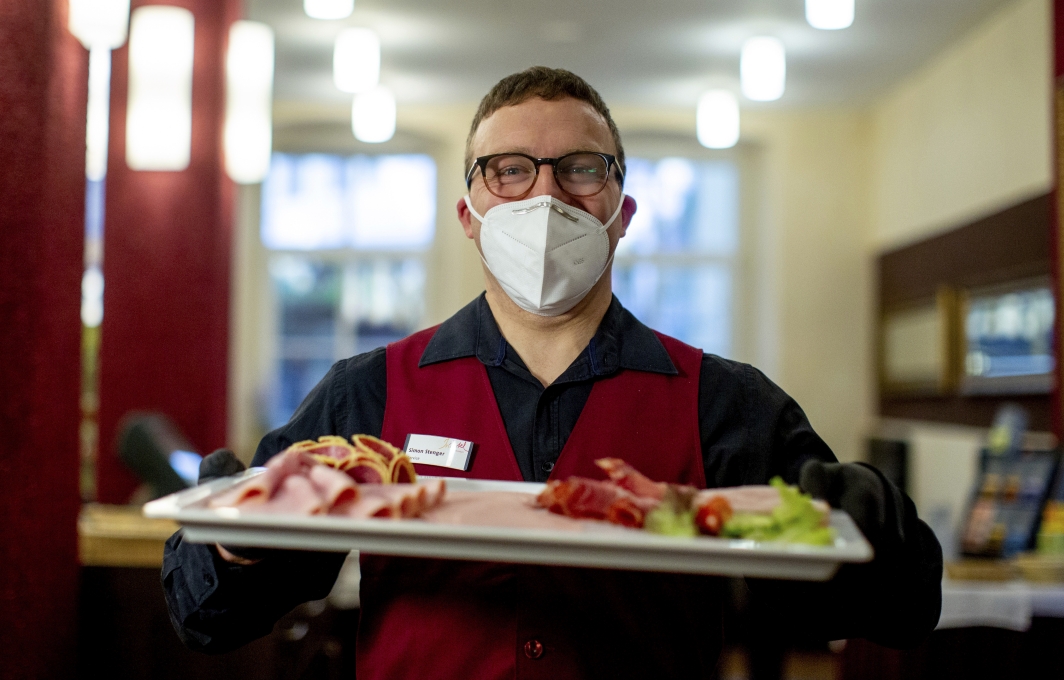 Ein Mann mit Mund-Nasen-Bedeckung und Hotelkleidung trägt ein Tablett mit Aufschnitt.