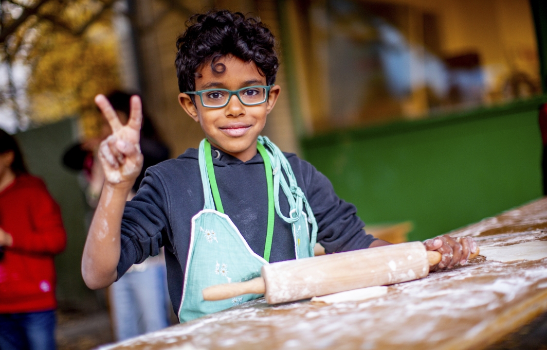 Ein Junge mit Brille rollt Pizzateig aus. Er lächelt und macht das Victory-Zeichen.