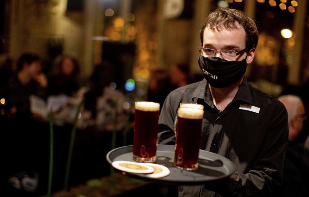 Ein Mann mit Mund-Nasen-Bedeckung trägt ein Tablett mit drei Biergläsern. Im Hintergrund ist der Gastraum der Schankhalle zu erkennen.