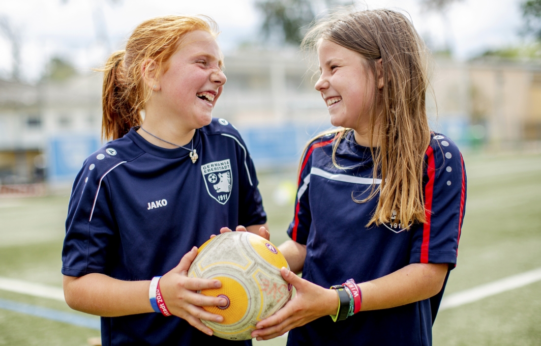 Zwei Mädchen halten gemeinsam einen Fußball in den Händen und lachen sich an.
