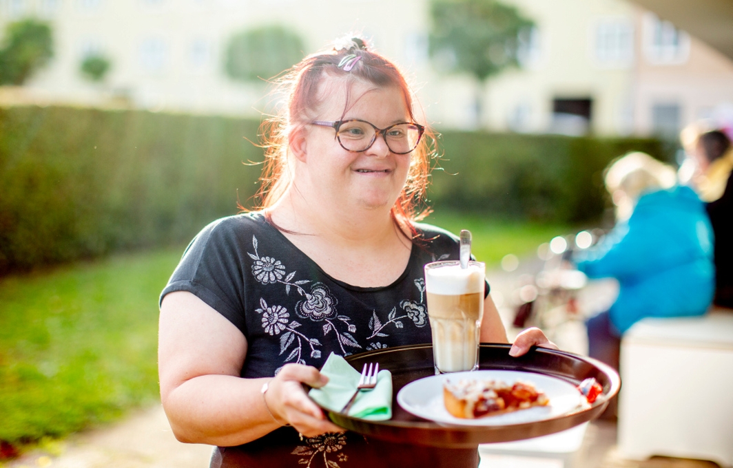 Eine Frau mit Down-Syndrom hält ein Tablett mit Kaffee und Kuchen.