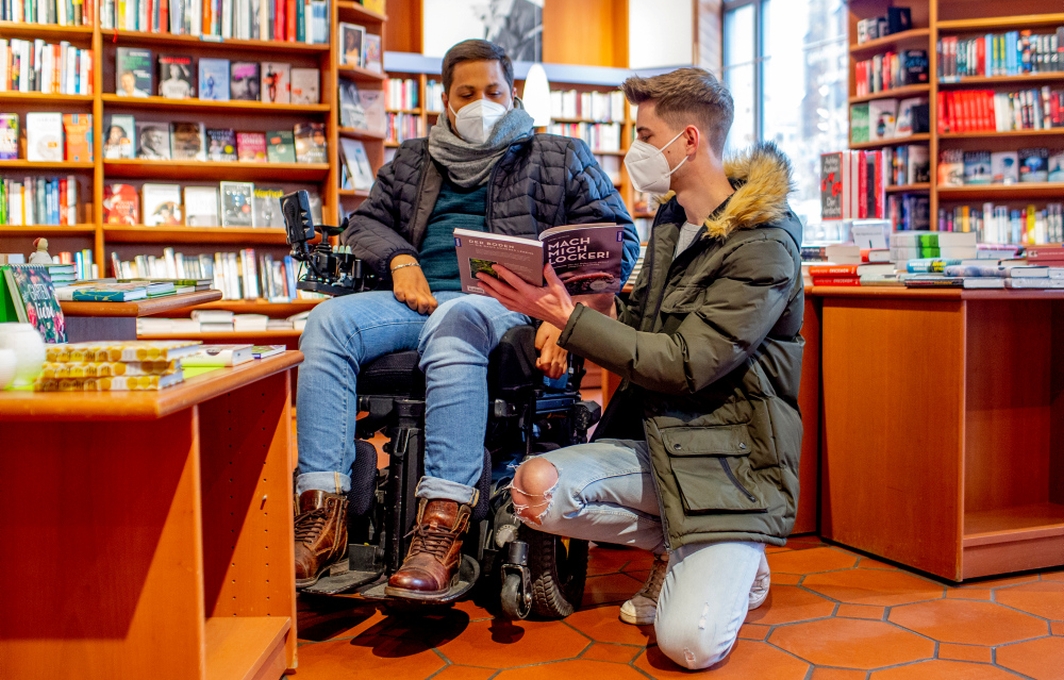 Ein Mann im elektrischen Rollstuhl und ein weiterer Mann besuchen eine Buchhandlung. Beide tragen Mund-Nase-Bedeckungen und schauen gemeinsam in ein Buch.