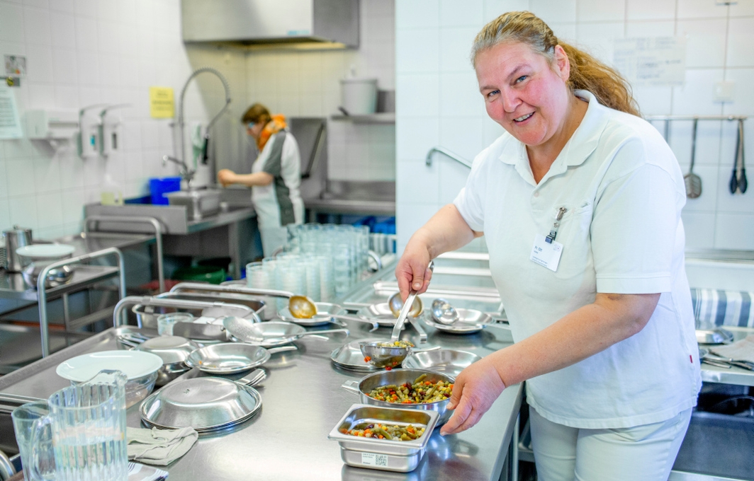 Ein Frau in weißer Kleidung steht in einer Großküche. Sie füllt mit einer Kelle buntes Gemüse in Schälchen.