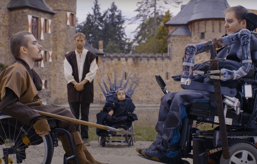 Zwei Männer in mittelalterlicher Kleidung und kampflustigen Blicken stehen einander zum Duell bereit gegenüber. Beide sitzen im Rollstuhl. Im Hintergrund ist eine Burg zu sehen.