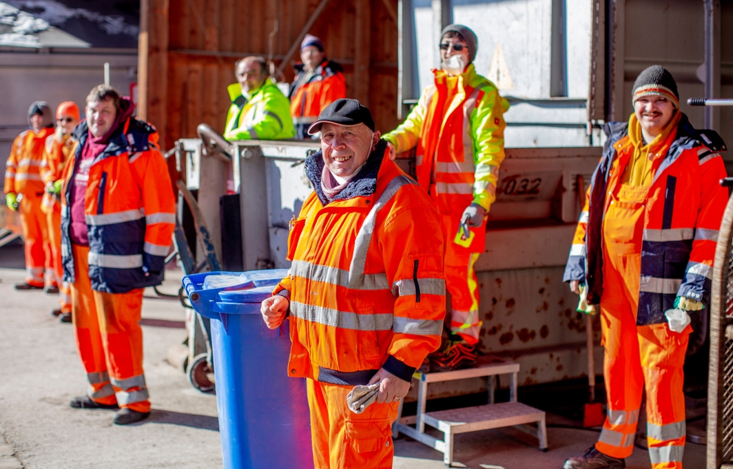 Eine Gruppe von Personen in orangefarbener Arbeitskleidung. Alle stehen auf Abstand zwischen Werststoff-Behältern und lächeln in die Kamera.