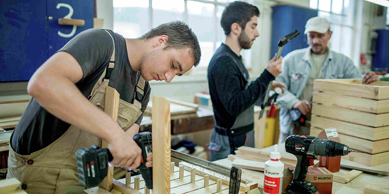 Ein junger Mann handerwerkelt, im Hintergrund stehen zwei andere Männer in der Werkstatt