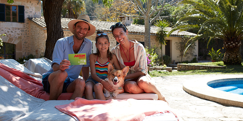 An einem Pool sitzt eine fröhliche Familie mit Hund. Der Vater hält das Glückslos in die Kamera.