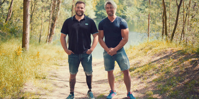 Zwei Männer, einer davon mit Prothese, stehen auf einer Waldlichtung.