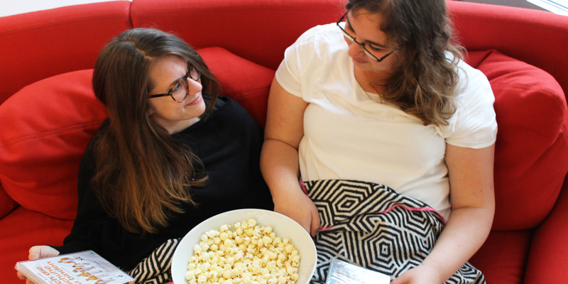 Zwei junge Frauen, die auf einer Couch sitzen. In der Hand haben sie verschiedene DVD-Hüllen und eine Schale Popcorn.