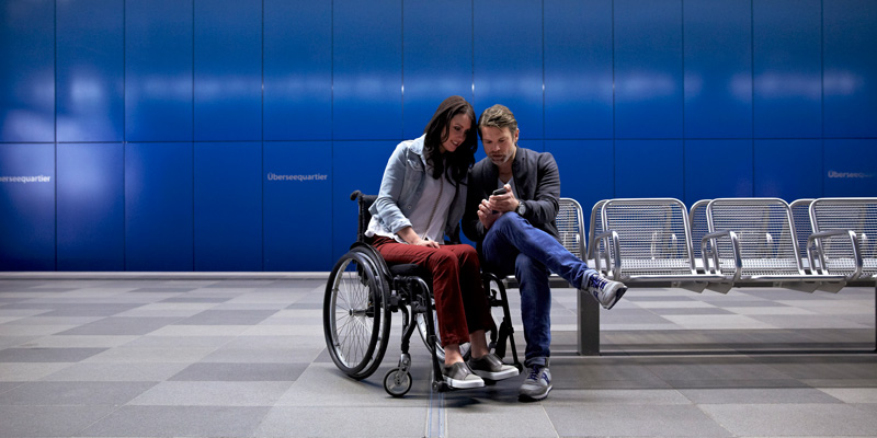 Eine Frau im Rollstuhl und ein Mann sitzen an einer Bahnhaltestelle und schauen auf ein Handy.