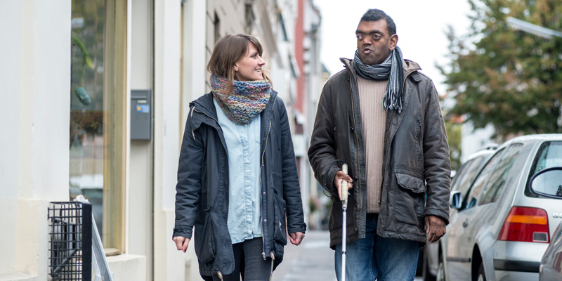 Eine Frau und ein Mann mit Blindenstock gehen über die Straße und lächeln sich an.
