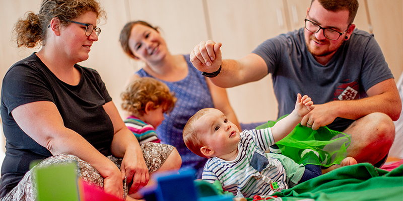 Eltern und ihre Kleinkinder sitzen in einem Raum zwischen Spielzeug und spielen mit ihren Kindern.