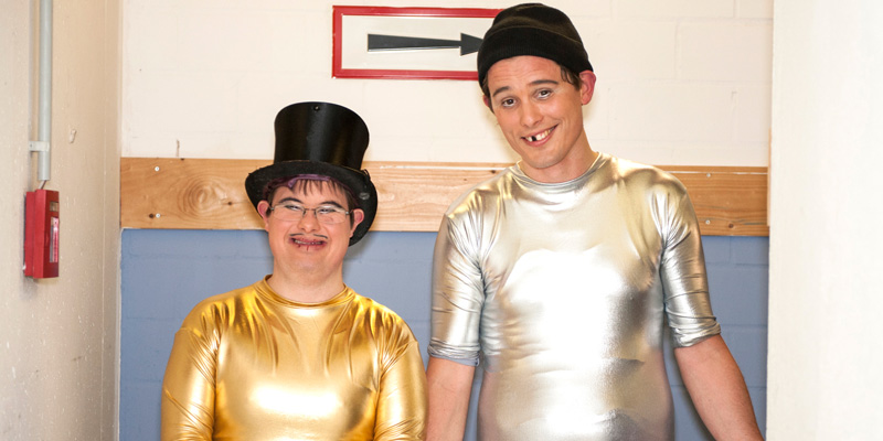 Ein Mann mit Down-Syndrom und ein Mann ohne Behinderung lächeln in die Kamera, sie tragen silber- und gold-glänzende Kleidung.