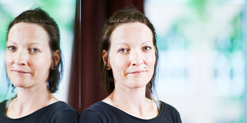 Das Gesicht einer Frau, die mit eindringlichem Blick in die Kamera schaut. Ihr Gesicht spiegelt sich in einer Glaswand, an der sie steht.