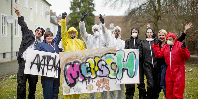 Menschen in Schutzkleidung halten Plakate hoch auf denen: Aktion Mensch gesprayt ist.