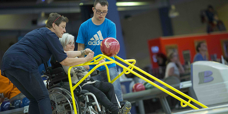 Rollstuhlfahrerin wird von einer Frau beim Einlegen der Bowlingkugel in ein Gestell unterstütztt