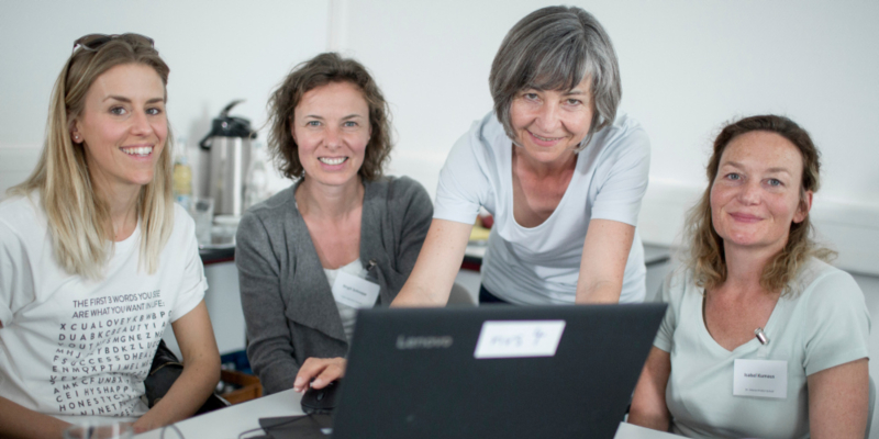 Vier Frauen sitzen in einem Lehrraum vor einem aufgeklappten Laptop und lächeln in die Kamera.
