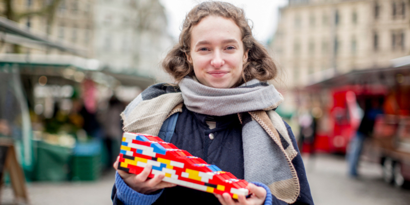 Eine junge Frau hält eine Legorampe in den Händen und lacht in die Kamera.