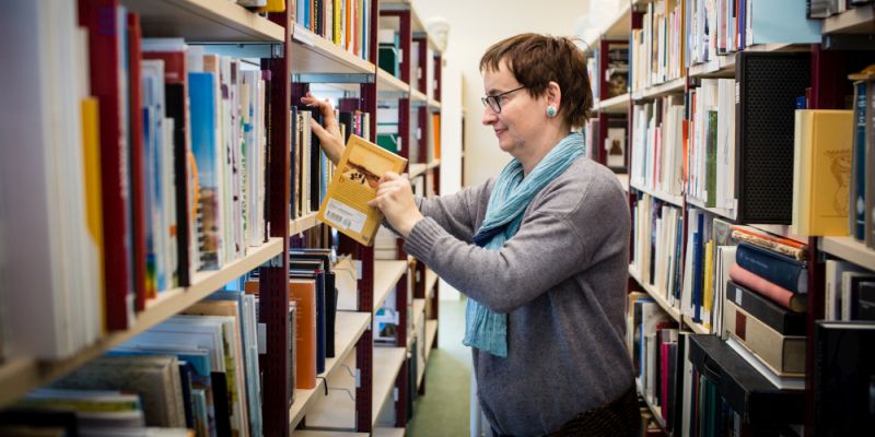 Eine Frau steht zwischen zwei Bücherregalen und sortiert Bücher.